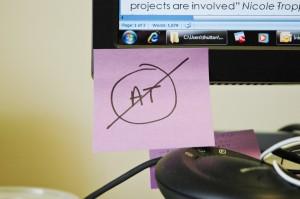 The word AT crossed out, written in marker on a sticky-note attached to a computer screen