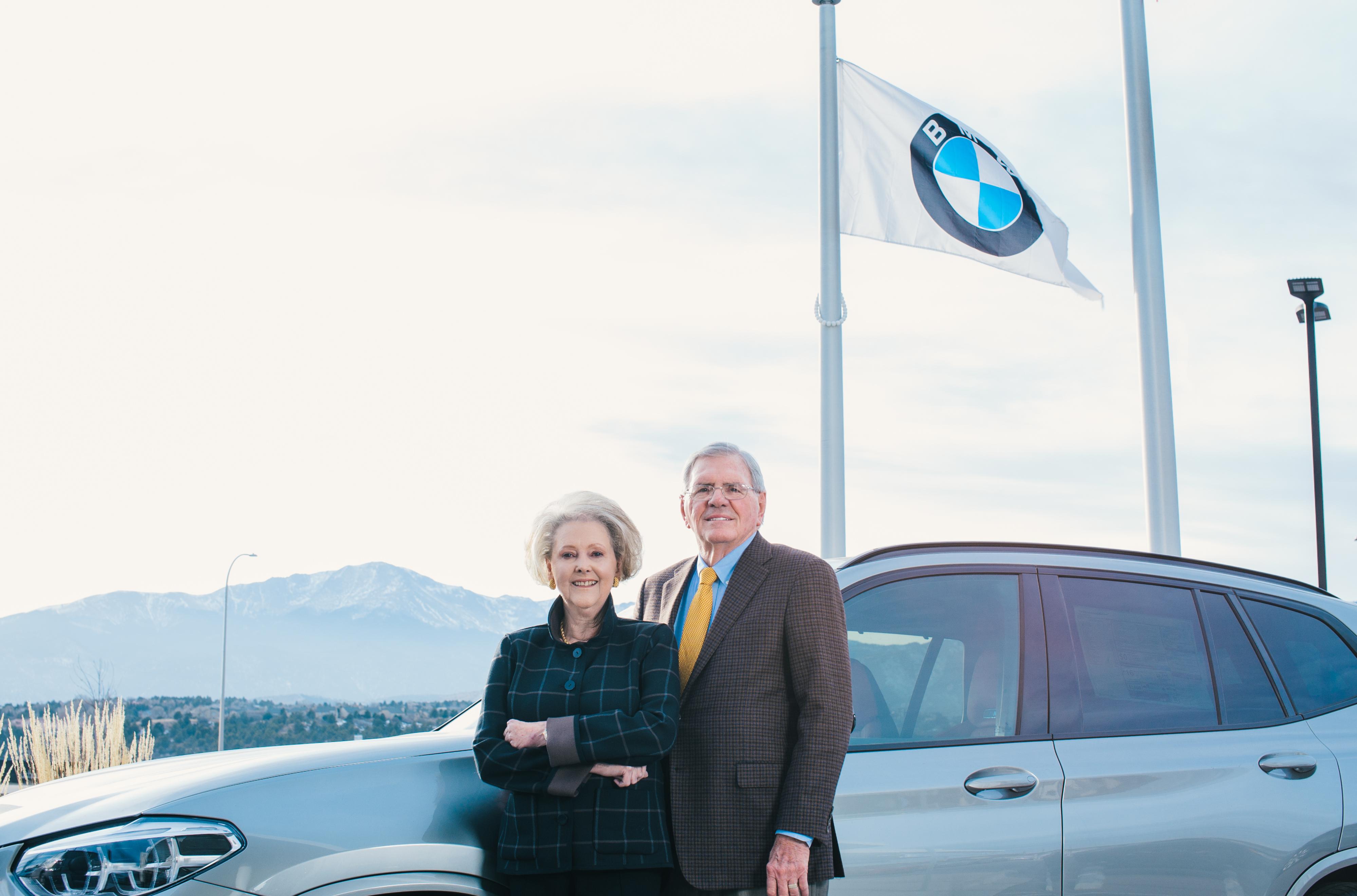 Phil and Ann Winslow standing in front of a car with a BMW flag and Pikes Peak in the background