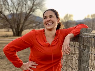 Professor Cerian Gibbes poses for a photo in a bright red blouse. She is leaning on a wood and wire fence in a field in winter.