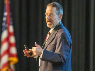 Chip Benight speaking at a conference with an American flag in the background
