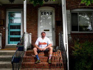 A person sits on his front step of his house with a face covering on