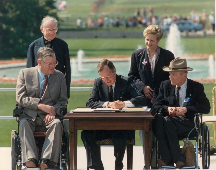 President George H. W. Bush sits at a table outside in the sunshine as he signs a document. He is flanked on either side by two men in suits sitting in wheelchairs. Two individuals look on. In the distance behind them is a fountain and a green lawn.
