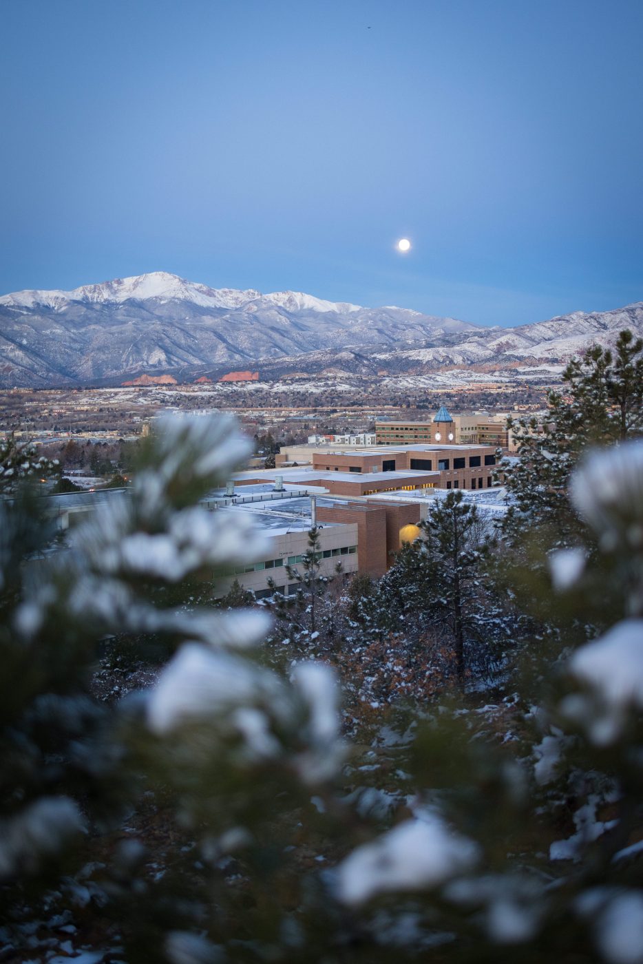 A snow-covered UCCS campus at sunrise in front of a full moon setting over Pikes Peak. Photo by Jeffrey M Foster